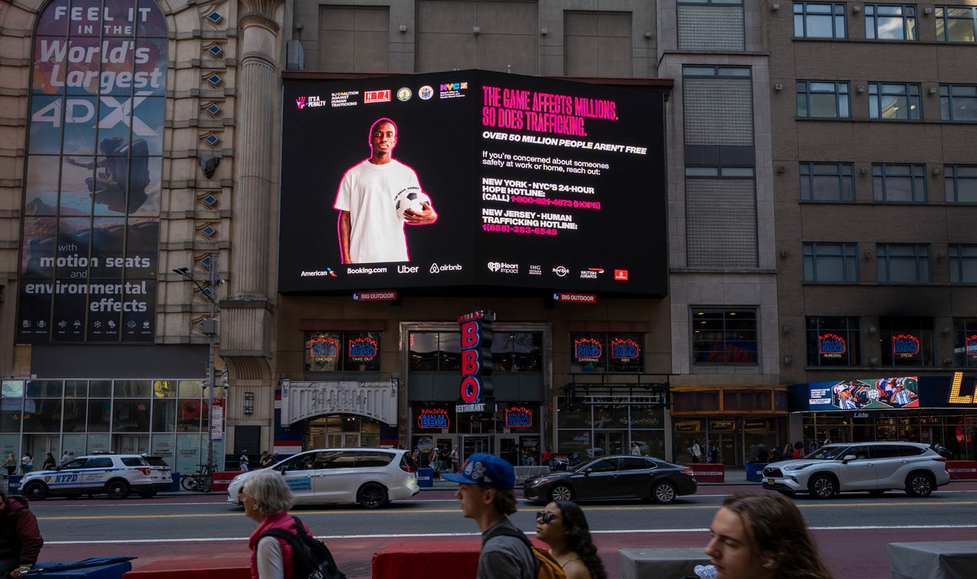 It's a Penalty campaign artwork on a large digital billboard in Times Square, New York, showing a footballer and the line 'The game affects millions. So does trafficking.'